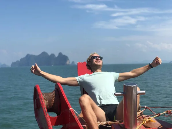 a blond guy in sunglasses and a t-shirt opened his hands to the sun on Board the ship on a hot day against the background of the sea and the Islands of Thailand, Phuket, enjoy life