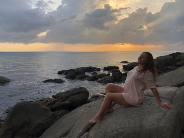 bir ışık plaj elbiseleri açık tenli kahverengi saçlı kız Karon Beach Phuket Tayland sahil şeridi taşlarının üzerinde oturur ve geç günbatımı hayran