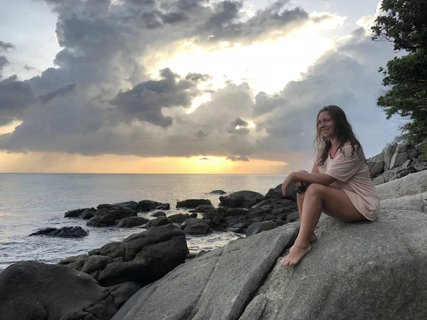 a light-skinned brown-haired girl in light beach clothes sits on the stones of the coastline of Karon beach in Phuket in Thailand and admires the late sunset