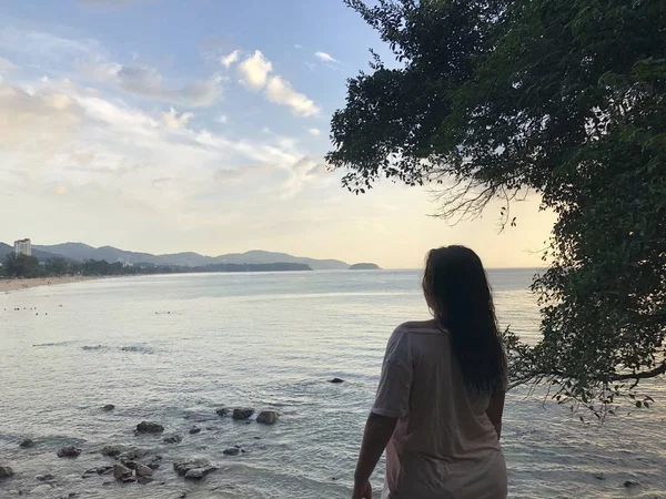 Une fille aux cheveux bruns à la peau claire en vêtements de plage légers est assise sur les pierres de la côte de la plage de Karon à Phuket en Thaïlande et admire le coucher du soleil tardif — Photo