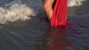 Köpüklü deniz dalgaları, uzun kırmızı elbise kenarlarına karşı kadın bacakları üzerinde yuvarlanıyor.