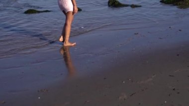 Çıplak ayaklı, pembe elbiseli genç bir kadın ıslak kumda yürüyor.