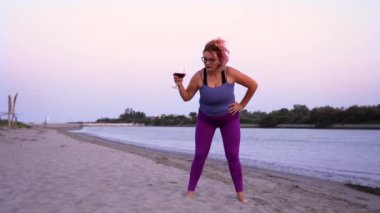 Komik tombul kadın, kumsalda egzersiz yaparken şarap içiyor.