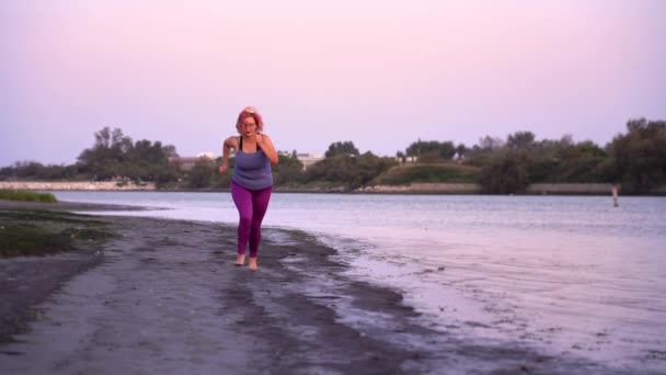 Συν-μεγέθους γυναίκα με γυαλιά τρέχει κατά μήκος άδεια παραλία του ποταμού — Αρχείο Βίντεο