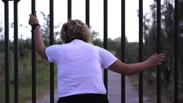 Молодая женщина в футболке осматривается возле металлических ворот — стоковое видео