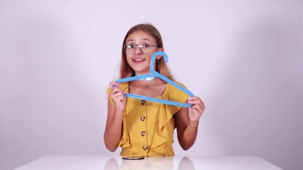Menina engraçada em blusa brinca com cabide sentado à mesa — Vídeo de Stock