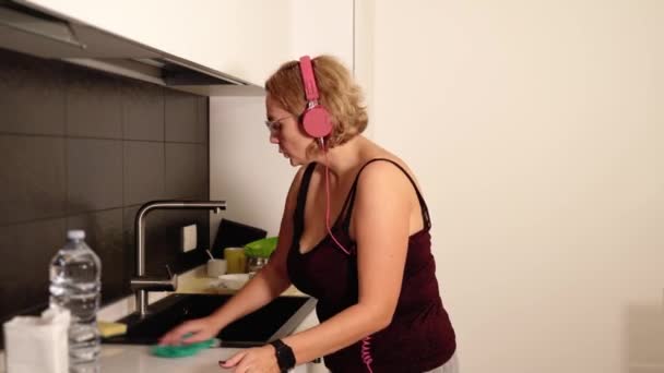 Nadwaga pani w słuchawkach tańczy i sprząta kuchnię — Wideo stockowe