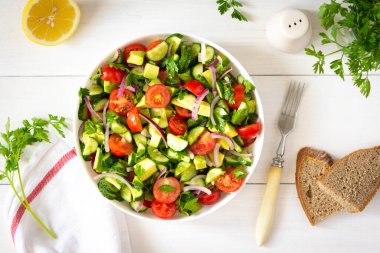 Taze olgun domates salatası, soğan, avokado, salatalık, maydanoz ve zeytin yağı, çatallı beyaz bir tabakta salata ve beyaz ahşap bir zeminde çay havlusu.