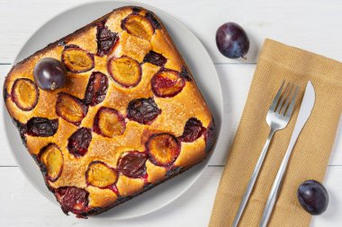 Beyaz ahşap arka planda yuvarlak bir tabakta lezzetli sulu erik turtası, kare pasta, peçetedeki çatal bıçak takımı, üst manzara.
