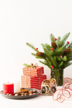 Hediye kutuları ve mumlar, tarçınlı kurutulmuş turunçgil ve fındık dilimleri, beyaz duvar arkasında bir vazoda köknar ağacı dalları ve koniler, yeni yıl ve noel konsepti.