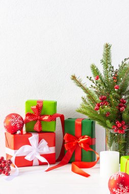 Kırmızı ve yeşil hediye kutuları ve mum dolu balolar, köknar ağacı dalları ve beyaz duvar arka planında bir vazoda şahin dikeni dalları, yeni yıl ve noel konsepti
