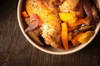 Koyu ahşap zemin üzerinde sebzeli fırında tavuk tabağı, patlıcanlı tavuk budu, balkabağı, soğan ve fırında pişmiş havuç.