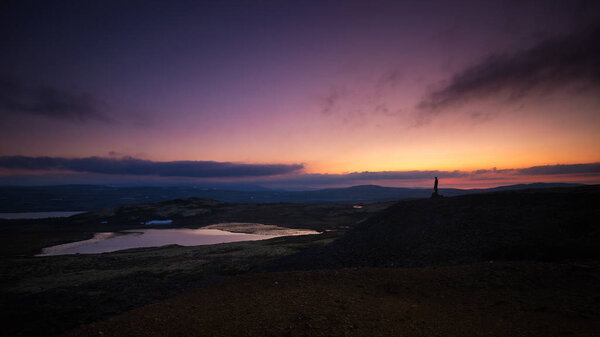 Distant silhouette of a man, standing on a mountain, watching midnight light. Nordgruvefeltet area in Norway.