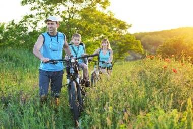 father with a bicycle goes through a meadow with poppies in a chair sitting his son. They enjoy nature. Outdoor sport activity.
