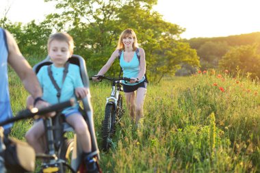 Mather with a bicycle goes through a meadow with poppies follows family. Woman enjoy nature. Outdoor sport activity.