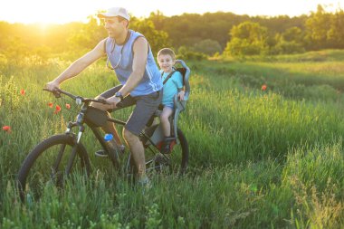Happy father and child in bicycle chair riding by the meadow in beautiful landscape. Healthy Lifestyle Concept.