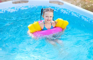 Küçük kız evinde yüzme havuzunda şişirilebilir daireler çiziyor, dışarıda eğleniyor. Çocuğa sarı yüzme yardımları