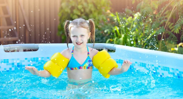 Little girl is splashing in frame swimming pool at home outdoor, having fun. Yellow swimming aids on child