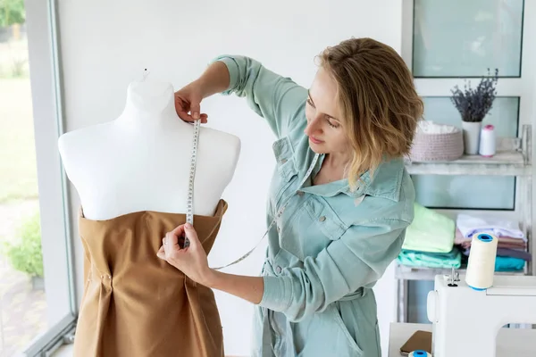 Female tailor or seamstress taking mannequin measurements for cloth pattern with measuring tape in fashion design studio, woman designer working with dummy in workshop