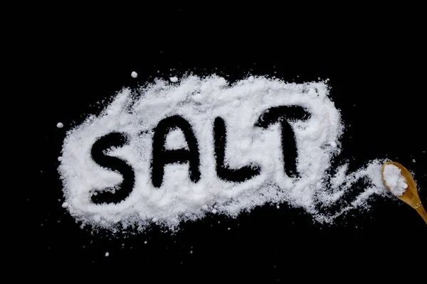 The word salt written into a pile of white salt and salt shaker on black background.