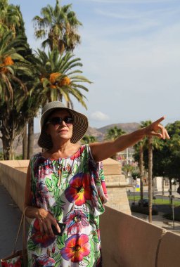 Orta yaşlı, şapkalı ve çiçekli elbiseli bir kadın İspanyol yoğunluğuyla cadde boyunca uzağa bakıyor.