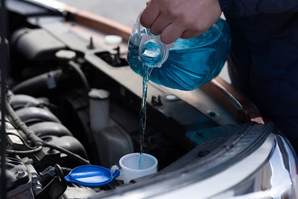 O homem está segurando garrafa de anticongelante azul nas mãos e derramando líquido anticongelante para lavar a tela do carro — Fotografia de Stock