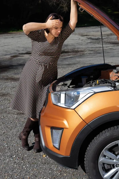 Desglose del coche. Mujer concentrada está sosteniendo la mano en la capucha del vehículo y mirando dentro de ella mientras está de pie al aire libre — Foto de Stock