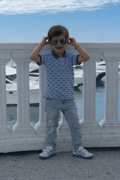 Portret van een modieuze jongen dragen zonnebril die genieten van een vakantie in de jachthaven. Hij draagt een blauwe jeans en een t-shirt — Stockfoto