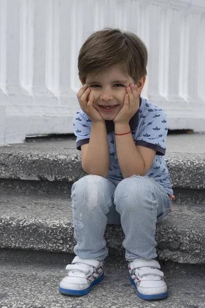 Joli garçon est assis sur les marches et s'ennuie. Un enfant à la mode portant un jean bleu et un t-shirt . — Photo