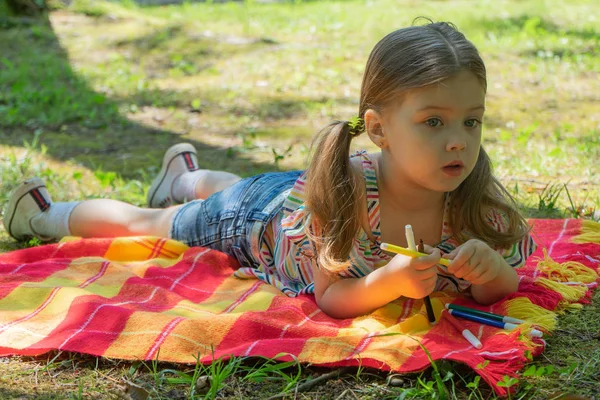 Petite fille montre feutre-pointe stylos colorés, Petite fille de trois à quatre ans se trouve sur une couverture lumineuse dans le jardin, elle tient dans sa main marqueurs colorés — Photo