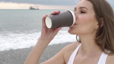 Gülümseyen sarışın kız sahilde bir karton camdan kahve içiyor, Deniz veya okyanus yanında gün batımında close-up