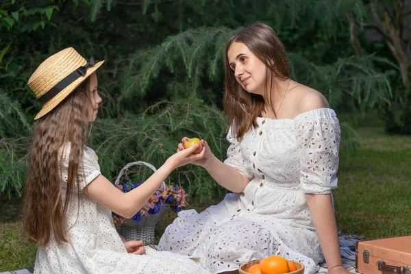 Vegetarian family on a picnic. Mother and daughter dressed in white dresses they sit on the plaid and eat fruits. Healthy lifestyle and nutrition