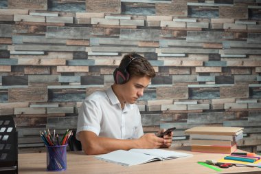 Young student sitting at desk in headphone and using his smartphone doing homework clipart