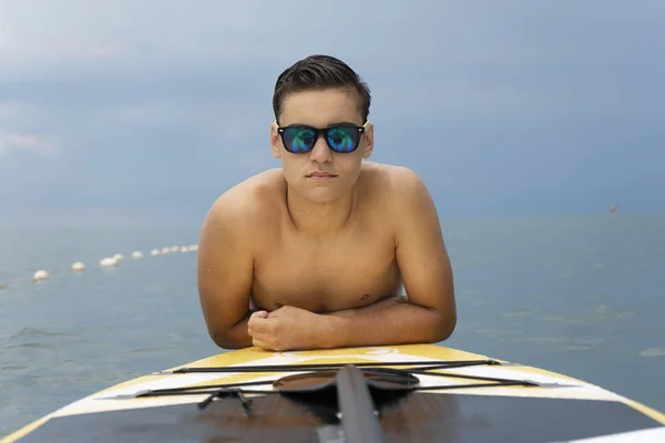 Modelo de moda em óculos de sol posando na água com prancha de surf sup. Esportes aquáticos, moda e estilo — Fotografia de Stock