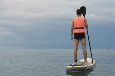 Denizin arka planında can yeleği giymiş sup board'da genç bir adam. Arka daki sörfçüden görüntüle