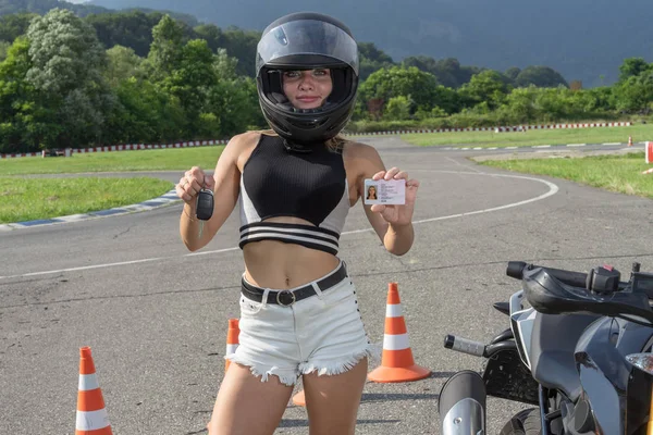 Concept of driving school for scooter. Girl standing next to a motorcycle. Woman in a helmet showing driving license and motorbikes key.