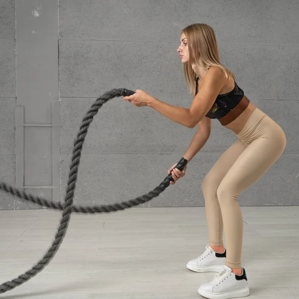 A girl training with battle rope in gym. Slim woman doing cardio load in gym. Athletic female doing some cross-training exercises with a rope in fitness club