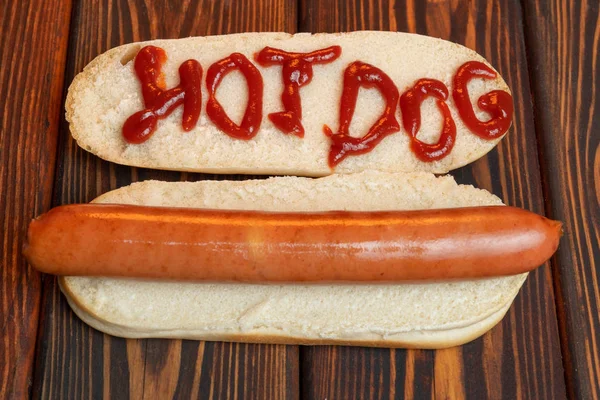 Hot dog bun with sausage on dark wooden background. On the bun is written in red ketchup - hot dogs — Stock Photo, Image