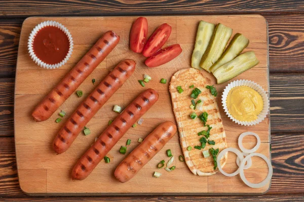 Sült kolbászok hagymával, savanyúságok, paradicsom, mustár, piros ketchup, fa asztallal. Grill — Stock Fotó