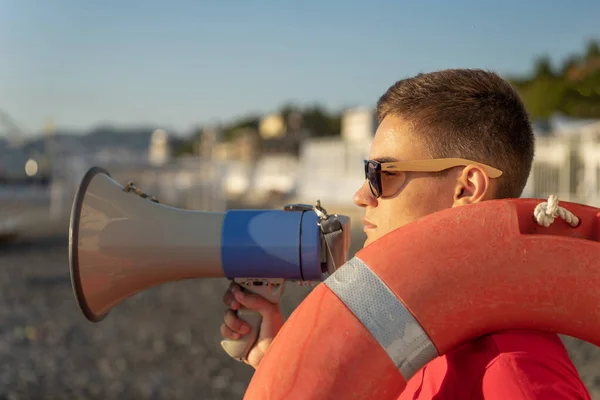 Młody ratownik z megafonem na tle plaży. Ratownik na służbie z boją ratunkową na plaży — Zdjęcie stockowe