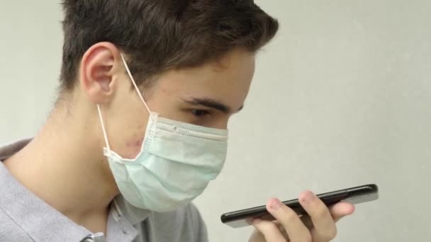 一个戴着一次性医疗保护面罩的年轻人在讲电话 使用虚拟数字语音识别辅助搜索在扬声器上保持青少年电话通话 — 图库视频影像