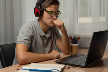 Evde dizüstü bilgisayar kullanan kulaklıklı yakışıklı bir genç. Çevrimiçi alışveriş, ev işi, serbest çalışma, çevrimiçi öğrenme, öğrenim kavramı. Uzaklık eğitimi