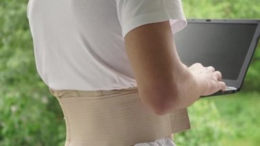 Bel desteği takan dizüstü bilgisayarı olan bir adam, doğanın arka planına karşı dizüstü bilgisayar kullanır. Parkta ortopedik korse giyen bir adam uzaktan çalışıyor..