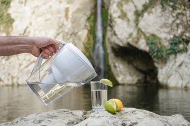 Su sürahisi. Bir adam suyu bir dağ nehrinin ve şelalenin arka planına karşı arıtmak için filtreyle sürahi kullanır.