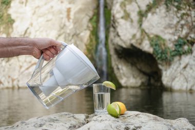 Water Pitcher. A man uses a jug with a filter to purify water against the background of a mountain river and waterfall clipart