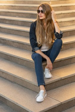 Uzun saçlı, kot takım elbiseli ve güneş gözlüklü bir sarışın mermer merdivenlerde oturuyor. Şehir ortamında merdivenlerde oturan bir kız.