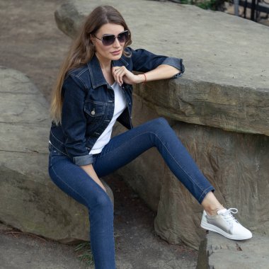 Kot pantolon giymiş şık bir sarışın şehir parkında bir kayanın üzerinde oturuyor. Kotlu bir kız ve taş bir masada beyaz bir tişört.