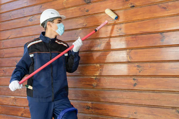 Koruyucu, tek kullanımlık bir maske ve eldivenle bir ahşap evin duvarını boyuyor.