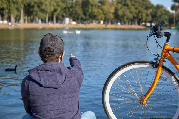 Young man with a cloth mask next to his bicycle pointing to a family of ducks on a lake in a park in Buenos Aires, Argentina, on a sunny winter afternoon during the outbreak of coronavirus, covid19