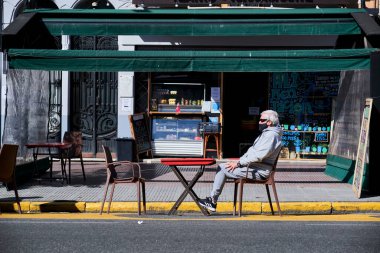 CABA, Buenos Aires / Argentina; Sept 19, 2020: senior man sitting in the sun at a table on the street, wearing a mask, during the coronavirus pandemic, covid-19 outbreak clipart
